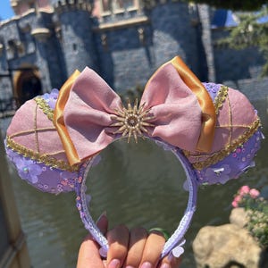 Peut inclure: Oreilles de Minnie Mouse violettes et roses avec un nœud rose et or et une broche en forme de soleil scintillante. Les oreilles sont décorées de paillettes et de petites fleurs violettes.