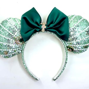May include: A sea-themed headband with seashell-shaped ears covered in light green sequins. A dark green satin bow is centered on the headband, adorned with a seahorse charm. The ears are embellished with pearls and decorative accents.