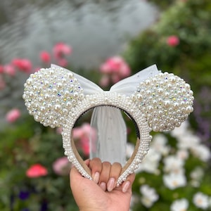 May include: White pearl and tulle Minnie Mouse ears headband. The ears are covered in white pearls and have a white tulle bow.