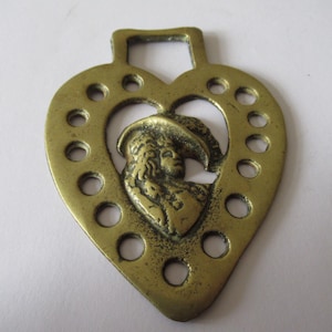 May include: A heart-shaped brass horse brass with a central cutout featuring a detailed relief of a person wearing a hat. The brass has a series of circular holes around the edge and a rectangular loop at the top.