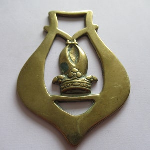 May include: A brass horse brass with a decorative design of a crown with a pointed top. The brass is a light gold color and has a worn finish.