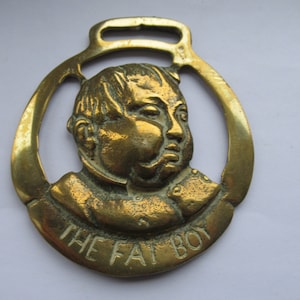 Op de afbeelding: Een messing paardenbrass met een reliëfbeeld van een mannengezicht en de tekst "The Fat Boy".