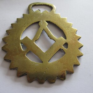 May include: A vintage brass horse brass with a sunburst design. The center features a square and compass symbol, with a loop at the top for hanging. The brass has a weathered appearance, suggesting age and use.