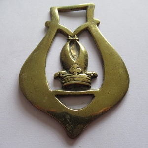 May include: A brass horse brass with a decorative design featuring a crown and a clover. The brass is a light gold color and has a shiny finish.