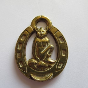 Könnte beinhalten: Ein goldfarbener Metall-Hufeisen-Anhänger mit einer kleinen, detaillierten Figur eines Kobolds, der darin sitzt. Der Kobold hält ein kleines Objekt in seiner Hand.