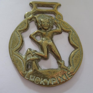 May include: Vintage brass horse brass featuring a pixie figure within a decorative oval frame. The pixie is depicted in a playful pose, with the words "LUCKY PIXIE" inscribed at the bottom. The brass has a loop at the top for hanging.