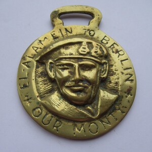 May include: A vintage brass medallion with a loop at the top. The medallion features a relief portrait of a man in a military cap, surrounded by the text "EL ALAMEIN TO BERLIN OUR MONTS".