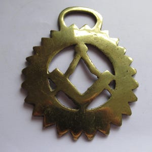 May include: A vintage brass horse brass with a sunburst design. The center features a diamond shape with a vertical line and a horizontal line. The brass has a loop at the top for hanging.