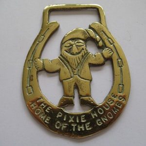 May include: A polished brass horse brass featuring a central figure of a gnome wearing a pointed hat, positioned within a horseshoe frame. The text THE PIXIE HOUSE THE HOME OF THE GNOMES is engraved along the bottom curve of the horseshoe.