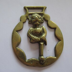 May include: A brass horse brass featuring a central figure of a seated monkey or creature within a circular frame with a scalloped edge. The object has a rectangular loop at the top for attachment and a smooth, metallic gold-toned finish.