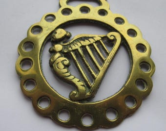 May include: A brass horse brass featuring a harp design within a circular frame. The frame has a scalloped edge with evenly spaced holes. The harp is detailed with decorative scrollwork.