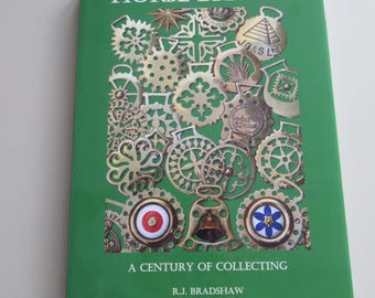 May include: A green book titled HORSE BRASSES A CENTURY OF COLLECTING by R.J. BRADSHAW. The cover features a collage of various brass horse brasses in circular, star, and bell shapes, including two pieces with red and blue floral centre accents.
