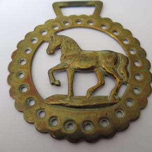 May include: A vintage brass horse brass featuring a horse figure. The horse is set within a circular frame with decorative holes. The brass has an aged, tarnished appearance, showing signs of wear. The horse is depicted in a walking pose.