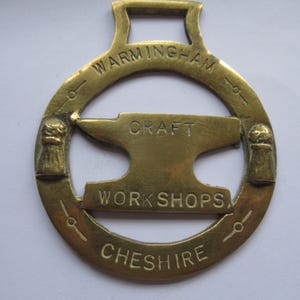Puede incluir: Un herraje de latón vintage con las palabras "WARMINGHAM CRAFT WORKSHOPS CHESHIRE" que rodean un diseño de yunque central. El latón tiene un lazo en la parte superior para colgar y elementos decorativos a cada lado.