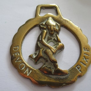 May include: A brass horse brass with a raised design of a pixie sitting on a rock. The brass is inscribed with the words "Devon Pixie".