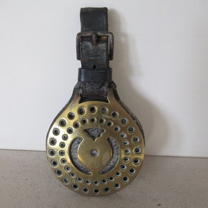 May include: A brass horse brass with a leather strap. The brass has a circular design with a heart-shaped cutout in the centre. There are small holes around the edge of the brass.