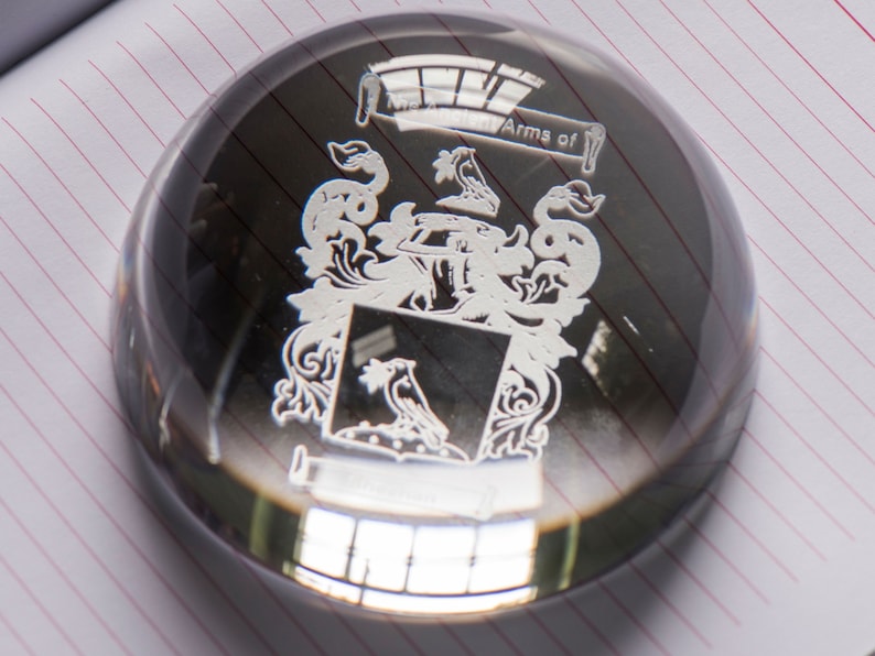 Peut inclure: Un presse-papier en verre transparent avec un motif grav&eacute; blanc d'un blason. Le motif comprend un &eacute;cu avec un oiseau, une couronne et des accents floraux. Le texte "Arms of" est grav&eacute; au-dessus de l'&eacute;cu.