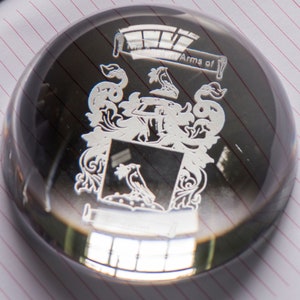 Peut inclure: Un presse-papier en verre transparent avec une gravure argent&eacute;e d'un blason. La gravure comprend un &eacute;cu avec un oiseau, une couronne et d'autres symboles h&eacute;raldiques. Le texte "Arms of" est &eacute;galement grav&eacute; sur le presse-papier.