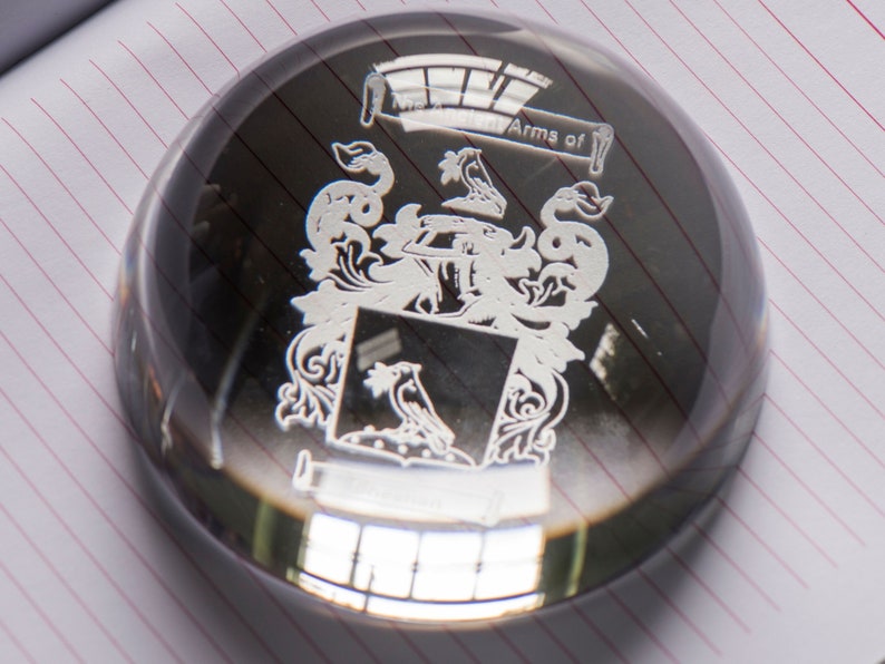 Peut inclure: Un presse-papier en verre transparent avec une gravure argent&eacute;e d'un blason. La gravure comprend un &eacute;cu avec un oiseau, une couronne et d'autres symboles h&eacute;raldiques. Le texte "Arms of" est &eacute;galement grav&eacute; sur le presse-papier.