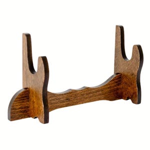 May include: A dark brown wooden display stand. It has two upright supports with curved tops and a wavy base. The stand is designed to hold an item. The stand is made of wood and has a varnished finish.