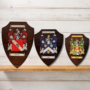 Op de afbeelding: Drie houten schilden met familiewapens en motto's. De schilden zijn bruin met gouden versieringen en tonen kleurrijke wapens met heraldische symbolen. De motto's zijn "John Dixon Bells Bells Dixon a Dixon", "Wees rechtvaardig en vrees niet" en "Ik sta vast in mijn orde".
