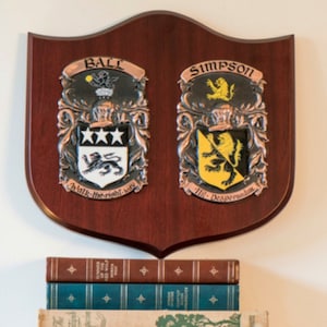 Puede incluir: Una placa de madera con dos escudos de armas. El escudo de armas de la izquierda es para la familia Ball y presenta un león negro con tres estrellas encima. El escudo de armas de la derecha es para la familia Simpson y presenta un león amarillo con un león negro encima. La placa está montada en una pared sobre una pila de libros.