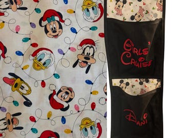 Op de afbeelding: Witte stof met een patroon van Mickey Mouse, Minnie Mouse, Donald Duck en Goofy met kerstmutsen en kerstverlichting. Een zwarte hangende organizer met vakken heeft de tekst "Girls Cruise", "Dani" en "Liv" in het rood.