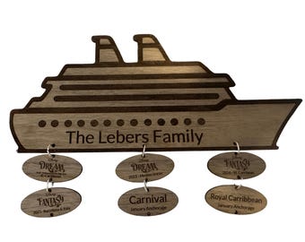 Könnte beinhalten: Ein hölzernes Familienurlaubsschild in Form eines Kreuzfahrtschiffs mit dem Text "The Lebers Family". Unter dem Schiff befinden sich sechs ovale Anhänger mit Kreuzfahrt-Details, darunter "DREAM", "FANTASY" und "Carnival".