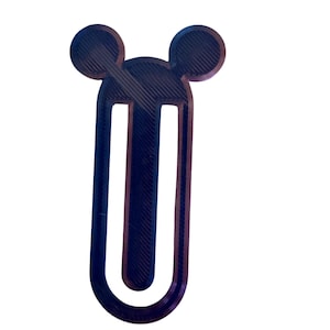 Puede incluir: Un marcapáginas azul oscuro con forma de cabeza de Mickey Mouse. El marcapáginas tiene forma de U con dos orejas circulares en la parte superior. El diseño está impreso en 3D y tiene líneas de capa.
