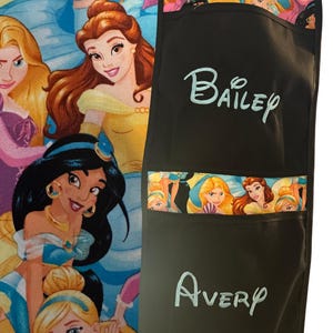 Puede incluir: Delantal negro con el nombre "Bailey" en escritura azul claro. El delantal tiene un bolsillo con un ribete de tela con temática de princesas. El nombre "Avery" está escrito en escritura azul claro en el bolsillo inferior. Un tejido colorido con temática de princesas es visible en el fondo.