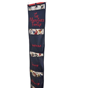 May include: A navy blue fabric organizer with multiple pockets, featuring the text "THE MARTINEZ FAMILY" in red script. Each pocket is labeled with a name in red, and the pockets have a Mickey Mouse pattern.