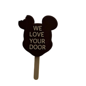Op de afbeelding: Een bruine chocolade Mickey Mouse-kop ijslolly op een stokje met de tekst "WE LOVE YOUR DOOR" gedrukt op de voorkant.