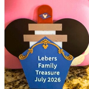 Puede incluir: Un letrero de tesoro familiar azul, dorado y blanco con el texto "Lebers Family Treasure July 2026". El letrero tiene forma de sombrero con orejas de Mickey Mouse y un icono de Mickey Mouse rojo y blanco.