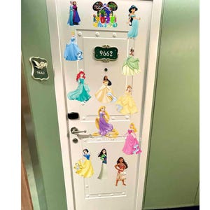 Pode incluir: Uma porta branca decorada com autocolantes de princesas. A porta tem uma maçaneta prateada e uma placa com o número do quarto. Os autocolantes apresentam personagens em vestidos coloridos, incluindo Cinderela, Ariel e Jasmine.