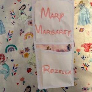 Puede incluir: Organizador de tela blanca con tres bolsillos, cada uno bordado con un nombre rosa: Mary, Margaret y Rozella. El organizador está sobre una tela estampada con ilustraciones de princesas y diseños florales.