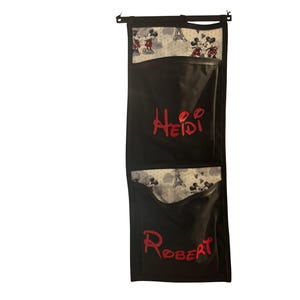 Puede incluir: Organizador colgante negro con dos bolsillos. La parte superior del organizador y la parte superior de cada bolsillo presentan una tela blanca con un estampado de Mickey y Minnie Mouse, y la Torre Eiffel. El nombre "Heidi" est&aacute; bordado en rojo en el bolsillo superior y "Robert" en el inferior.