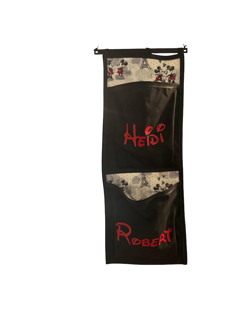 Puede incluir: Organizador colgante negro con dos bolsillos. La parte superior del organizador y la parte superior de cada bolsillo presentan una tela blanca con un estampado de Mickey y Minnie Mouse, y la Torre Eiffel. El nombre "Heidi" est&aacute; bordado en rojo en el bolsillo superior y "Robert" en el inferior.
