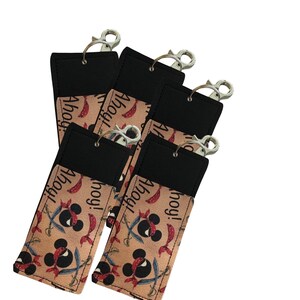 May include: A collection of six black and tan lip balm holders with silver-colored clasps. The holders feature a pirate-themed design with a Mickey Mouse-like skull and crossbones, swords, and the word "Ahoy!".