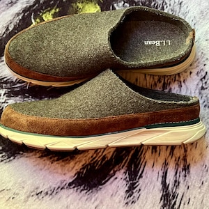 May include: A pair of olive green felt slip-on shoes with brown suede trim along the base. The shoes feature a white athletic-style midsole with a thin teal accent stripe and the L.L.Bean logo printed on the inner footbed and embossed on the heel.