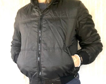 May include: A black puffer jacket with a zipper closure and a gold logo on the bottom. The jacket has a hood and a quilted design.