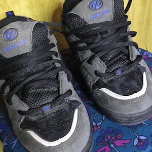 Puede incluir: Un par de zapatos Heely's grises y negros con detalles azules y cordones negros. Los zapatos tienen una franja blanca alrededor de la parte inferior del zapato.