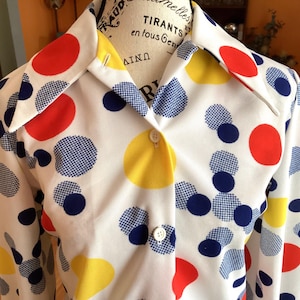 May include: White button-down shirt with a colorful polka dot pattern. The shirt features large red, yellow, and blue polka dots, with some blue dots having a checkered design. The collar and cuffs are white.