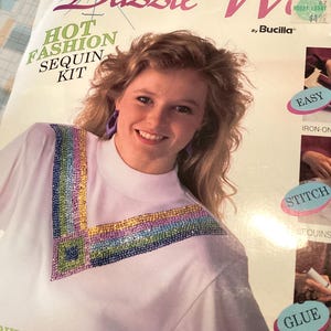 May include: A white t-shirt with a sequined design. The shirt features a V-neck design with a rainbow-colored sequin pattern. The packaging reads "Hot Fashion Sequin Kit" and "Dazzle W".