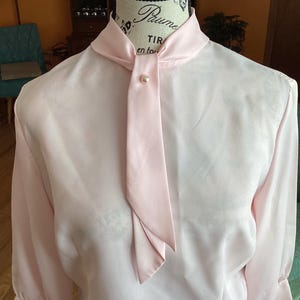 May include: A light pink blouse with a long tie that has a pearl button at the top. The blouse has long sleeves and a high collar.