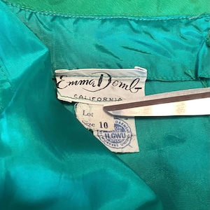 以下が含まれることがあります： ヴィンテージのティールグリーンの衣服タグのクローズアップ。ブランド名「Emma Domb」と「California」が印刷されています。タグにはサイズ10と組合ラベルも表示されています。
