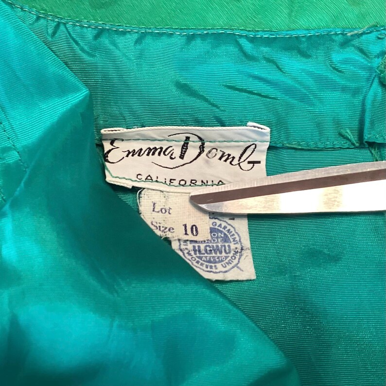 以下が含まれることがあります： ヴィンテージのティールグリーンの衣服タグのクローズアップ。ブランド名「Emma Domb」と「California」が印刷されています。タグにはサイズ10と組合ラベルも表示されています。