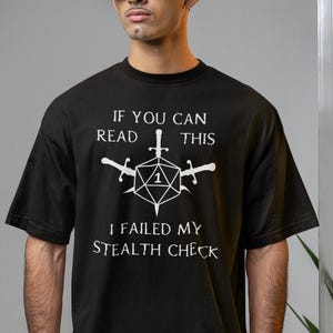 May include: A black t-shirt with a white graphic that reads "If you can read this I failed my stealth check". The graphic features a white dice with a 1 on it and three swords.