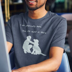 Puede incluir: Un hombre con una camiseta gris oscuro con el texto "Estoy físicamente aquí pero mi mente está ahí" y una silueta de una persona con un perro. Está sentado en un escritorio y mirando la pantalla de un ordenador.