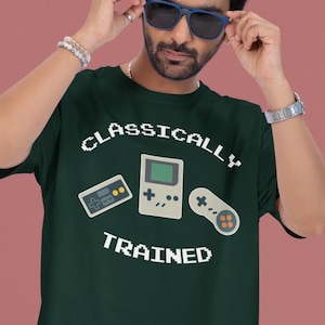 May include: Dark green t-shirt with the text "CLASSICALLY TRAINED" and pixelated images of a handheld game console and controller. The person is wearing blue sunglasses and denim shorts.