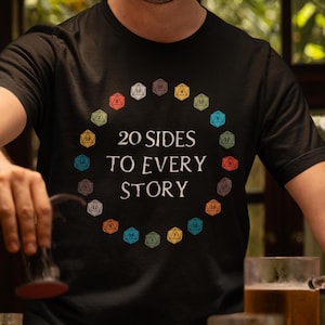 以下が含まれることがあります： 20面ダイスがカラフルな円形で描かれた黒いTシャツを着た人。Tシャツには「20 SIDES TO EVERY STORY」という文字が書かれている。